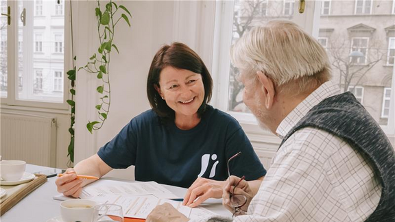 Eine lächelnde FSW-Mitarbeiterin sitzt an einem Tisch mit einem älteren Mann und berät ihn.