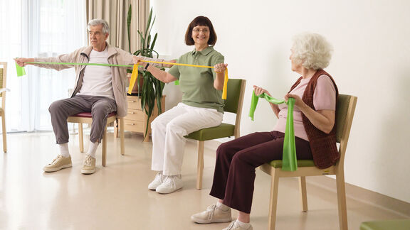 Eine FSW-Mitarbeiterin macht mit zwei Senior:innen Gymnastik mit Bändern in einem Tageszentrum für Senior:innen