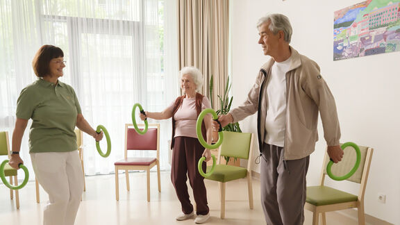 Eine FSW-Mitarbeiterin macht mit zwei Senior:innen in einem Tageszentrum Gymnastik mit Ringen