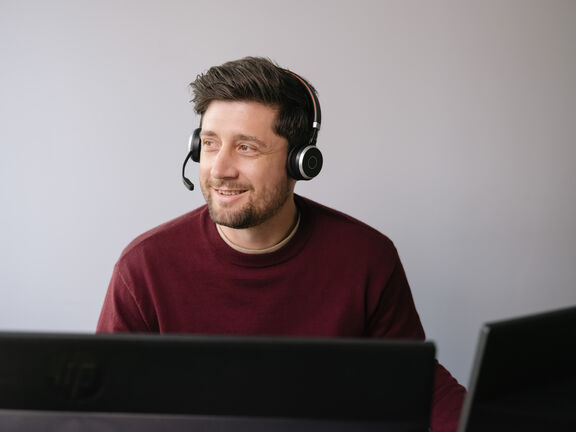 Ein junger Mann trägt ein Headset, sitzt auf einem Arbeitsplatz und blickt nach links
