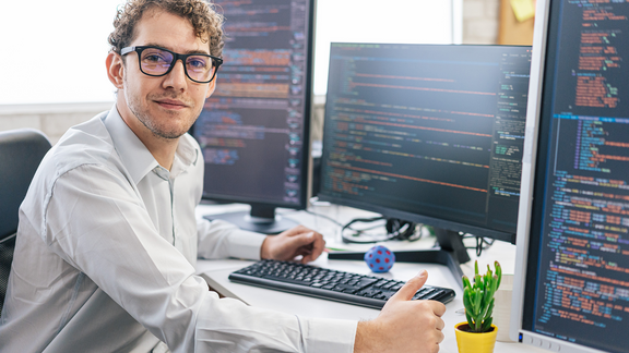 Ein junger Mann mit schwarzer Brille sitzt auf einem Schreibtisch mit drei Monitoren, die Code zeigen