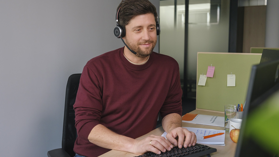 Ein junger Mann mit Headset sitzt vor einem Computer und lächelt