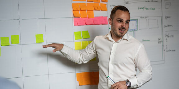 Ein junger Mann mit dunkler Hautfarbe steht vor einem Projektplan mit vielen Post-Its und erklärt