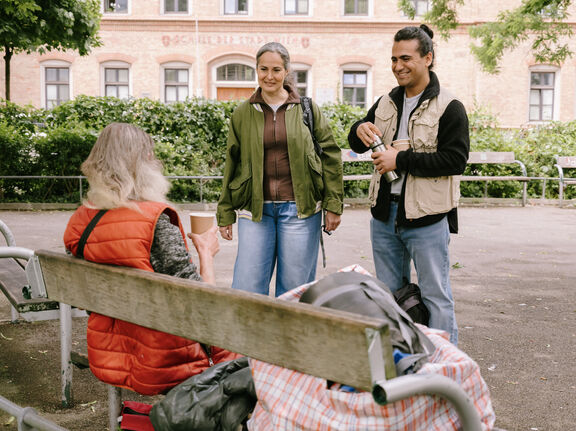 Ein FSW-Mitarbeiter und eine FSW-Mitarbeiterin sprechen im Park mit einer Person, die auf der Parkbank sitzt.