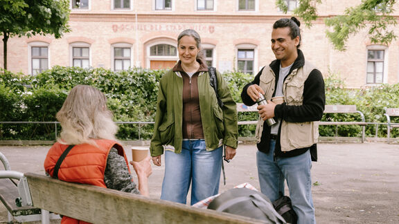 Ein FSW-Mitarbeiter und eine FSW-Mitarbeiterin sprechen im Park mit einer Person, die auf der Parkbank sitzt.