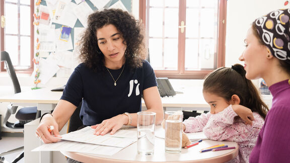 Eine FSW-Mitarbeiterin mit braunen Locken berät eine Frau mit Kopftuch und ihrer Tochter an einem Schreibtisch