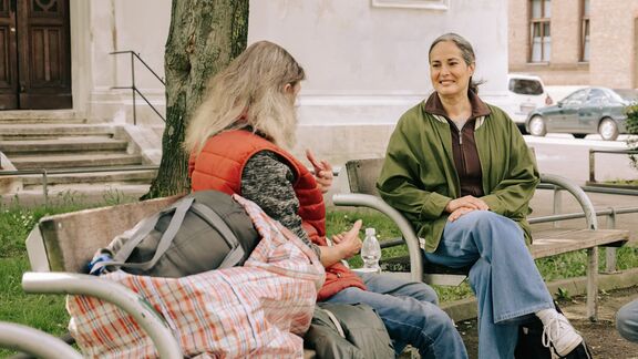 Eine Person mit weißen Haaren sitzt auf einer Parkbank und spricht mit einer FSW-Mitarbeiterin