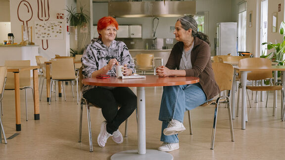 Eine FSW-Peer-Mitarbeiterin sitzt an einem Esstisch mit einer Frau mit orangenen Haaren