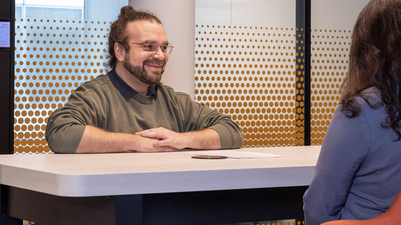 Ein Mann mit Bart und Brille sitzt lächelnd an einem Bürotisch und führt ein Gespräch mit einer Frau, die ihm gegenüber sitzt.