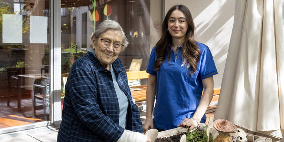 Eine lächelnde junge Mitarbeiterin im Freiwilligen Sozialen Jahr steht neben einer Seniorin.