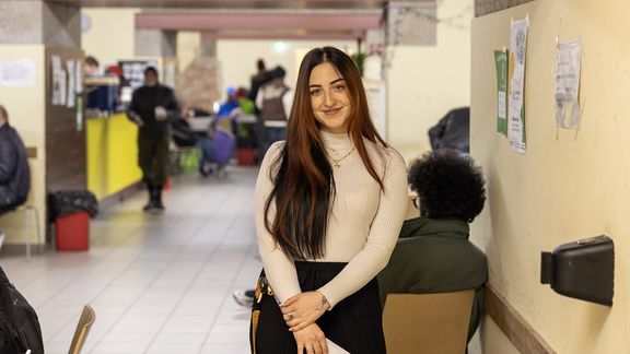 Eine lächelnde junge Frau mit langen dunklen Haaren steht in einem hellen, belebten Gemeinschaftsraum in einem Tageszentrum.