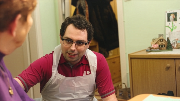 Ein lächelnder Pfleger mit Brille in einem Wohnzimmer im Gespräch mit einer älteren Dame.