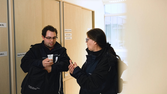 Zwei Pflegekräfte in dunkler Dienstkleidung stehen vor Materialschränken und besprechen die Anwendung eines Pflegeprodukts.