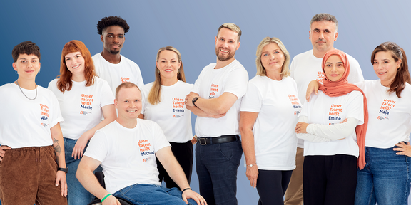 Zehn lächelnde Mitarbeiter des Fonds Soziales Wien in weißen T-Shirts vor hellblauem Hintergrund.