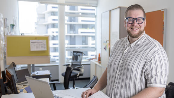 Ein lächelnder Mitarbeiter mit Brille arbeitet an einem Stehschreibtisch in einem hellen, modernen Büro des Fonds Soziales Wien (FSW).