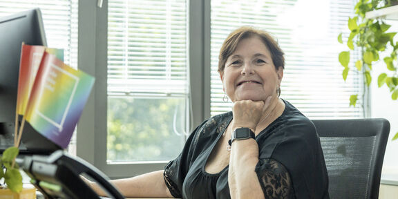 Eine lächelnde Frau mittleren Alters stützt ihr Kinn auf die Hand und blickt in die Kamera.