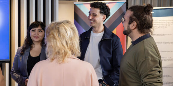 Vier lächelnde Kollegen stehen in einem modernen Büro zusammen und unterhalten sich.